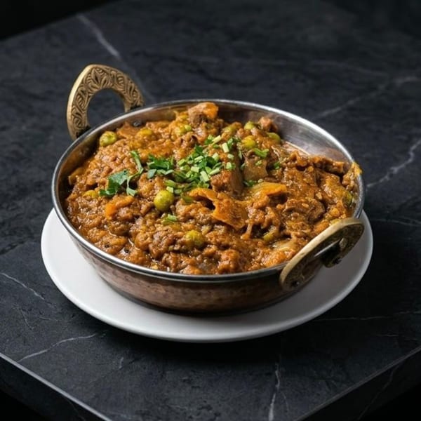 A copper karahi bowl filled with a rich, brown curry, garnished with green peas and fresh herbs.
