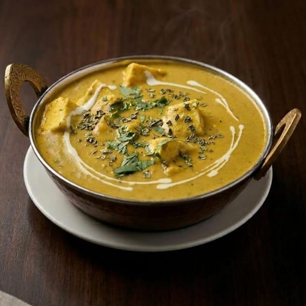 Steaming Indian curry with paneer, garnished with herbs and cream swirls, in a brass bowl.